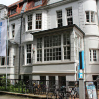 Das Bild zeigt die Fassade eines historischen, weißen Gebäudes mit großen Fenstern und einem Eingangsbereich. Vor dem Gebäude hängen zwei Fahnen mit der Aufschrift Innere Mission. Im Vordergrund sind Fahrräder abgestellt und eine kleine Treppe führt zum Eingang.